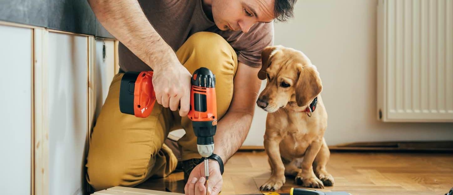 Renoveringslån – Låna pengar till renovering 6 Man som utför renovering, potentiell mottagare av ett renoveringslån, borrar i en planka med sin hund bredvid.